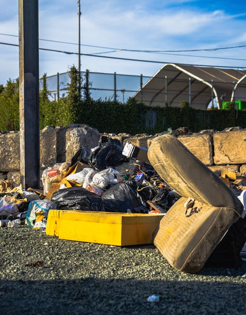 A pile of unmanaged rubbish on a gravel surface, consisting of black plastic trash bags, discarded cardboard and paper, and a worn, dirty beige car seat that appears to have been abandoned. The refuse is positioned in front of a low stone wall made of irregularly shaped, weathered stones, with some debris spilling onto the ground. In the background, a tall metal utility pole with attached wires extends upward, and behind the refuse, there are green shrubs and a hedge. Further back, a large, curved metal structure or tent with a fabric roof can be seen, along with a clear blue sky and some distant trees, suggesting an open outdoor area. The scene is illuminated by natural daylight, emphasizing the contrast between the clutter and the surrounding environment. This image depicts unmanaged waste often associated with private rubbish removal services, such as those offered by Rubbish Removal Battersea, and highlights the importance of proper disposal to prevent illegal dumping.