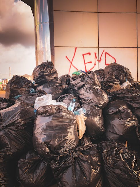 A pile of unmanaged rubbish on a gravel surface, consisting of black plastic trash bags, discarded cardboard and paper, and a worn, dirty beige car seat that appears to have been abandoned. The refuse is positioned in front of a low stone wall made of irregularly shaped, weathered stones, with some debris spilling onto the ground. In the background, a tall metal utility pole with attached wires extends upward, and behind the refuse, there are green shrubs and a hedge. Further back, a large, curved metal structure or tent with a fabric roof can be seen, along with a clear blue sky and some distant trees, suggesting an open outdoor area. The scene is illuminated by natural daylight, emphasizing the contrast between the clutter and the surrounding environment. This image depicts unmanaged waste often associated with private rubbish removal services, such as those offered by Rubbish Removal Battersea, and highlights the importance of proper disposal to prevent illegal dumping.