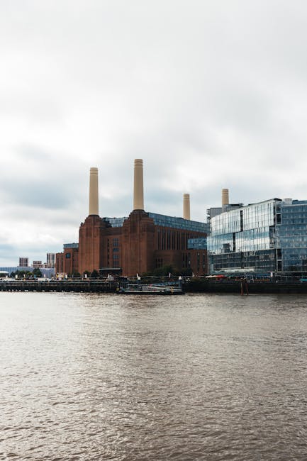 A large industrial building with a reddish-brown brick exterior and four tall beige chimney stacks is situated along a riverbank, with the river in the foreground showing slight ripples on its surface. Adjacent to the building, to the right, there is a modern glass structure with reflective windows, some of which are tinted dark, creating a sleek contrast with the industrial aesthetic. The scene is captured during cloudy daylight, with an overcast sky and diffuse light casting minimal shadows, highlighting the textures of the brickwork and glass. The setting appears urban, with boats moored along the river and a small section of a paved walkway or quay visible at the bottom of the image. The environment suggests an area undergoing redevelopment or repurposing, with Rubbish Removal Battersea occasionally involved in local waste management or site clearance for such industrial or mixed-use properties, supporting the context of private waste disposal or alternative rubbish collection methods in the Battersea area.