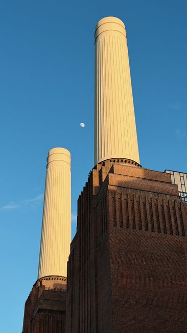 A close-up view of the upper section of Battersea Power Station, featuring two tall, cream-colored chimney stacks with vertical ridges and rounded tops, set against a clear blue sky. The larger chimney is positioned to the right, while the smaller chimney is slightly to the left. The base of the chimneys displays dark brown brickwork with textured detailing, indicating the historic industrial architecture. A small, bright full moon is visible in the sky near the larger chimney, adding a natural element to the scene. This image emphasizes the scale and structure of the power station, highlighting its distinctive design and the surrounding open sky, aligning with themes of site clearance and structural clearance in rubbish removal activities managed by Rubbish Removal Battersea.