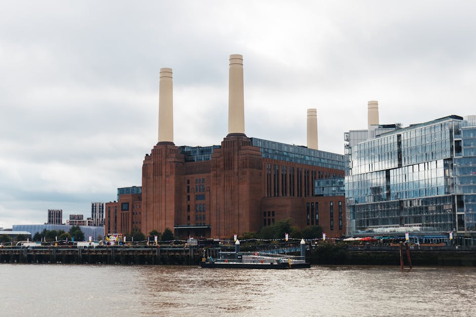 A large industrial building with a reddish-brown brick exterior and four tall beige chimney stacks is situated along a riverbank, with the river in the foreground showing slight ripples on its surface. Adjacent to the building, to the right, there is a modern glass structure with reflective windows, some of which are tinted dark, creating a sleek contrast with the industrial aesthetic. The scene is captured during cloudy daylight, with an overcast sky and diffuse light casting minimal shadows, highlighting the textures of the brickwork and glass. The setting appears urban, with boats moored along the river and a small section of a paved walkway or quay visible at the bottom of the image. The environment suggests an area undergoing redevelopment or repurposing, with Rubbish Removal Battersea occasionally involved in local waste management or site clearance for such industrial or mixed-use properties, supporting the context of private waste disposal or alternative rubbish collection methods in the Battersea area.