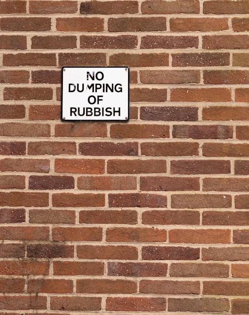 A rectangular white metal sign with black text reading 'NO DUMPING OF RUBBISH' is mounted on a red brick wall. The bricks are uniformly laid with light-colored mortar joints, demonstrating a typical brickwork pattern. The wall appears to be part of an outdoor setting, possibly a building facade or boundary wall, with no additional objects or environmental features visible in the image. The sign is positioned centrally within the frame, and the lighting suggests natural daylight, illuminating the brick surface evenly. The image conveys a clear message to prevent rubbish dumping, aligning with private waste management practices such as independent rubbish removal or site clearance services provided by companies like Rubbish Removal Battersea, although no specific waste removal activity is depicted in the scene.