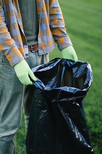 A person wearing a plaid shirt in shades of yellow, grey, and blue over a green top, with grey trousers and a brown belt, is holding open a large black rubbish bag with both hands, which is lined with a shiny black plastic material. The person is standing outdoors on a grassy area, with a blurred background of green lawn. They are wearing bright green gloves, likely for waste handling. The scene depicts an activity related to rubbish collection or waste disposal, aligning with services such as independent rubbish removal or on-site clearance offered by Rubbish Removal Battersea, emphasizing clean-up tasks involving bulky or general waste items.