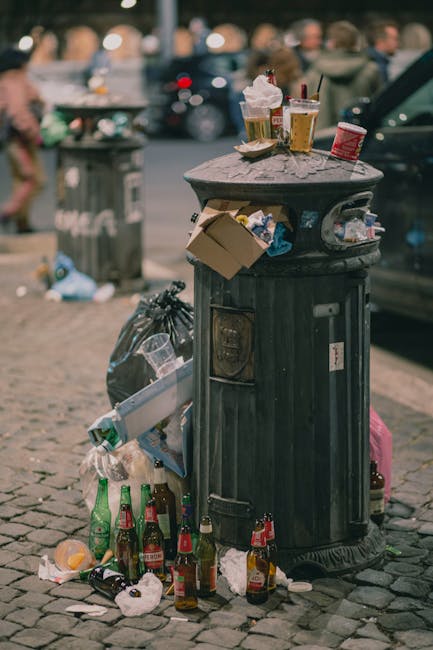 A black, cylindrical public rubbish bin located on a cobblestone sidewalk in an urban setting. The bin has a domed lid with an open slot for waste and is overflowing with various litter, including paper cups, plastic food containers, and a cardboard box, some of which have been placed on top of the lid. Around the base of the bin, there are multiple discarded glass beer bottles, some crushed or partly broken, as well as crumpled paper and plastic waste. In the background, a grey trash bag leans against the bin, and blurred figures of pedestrians and parked vehicles are visible, indicating a busy street environment. The scene is lit with natural daylight, capturing typical city littering and waste disposal challenges often seen in areas without immediate rubbish collection, illustrating the importance of private waste management services like those provided by Rubbish Removal Battersea for effective on-site or alternative rubbish handling in urban locations.